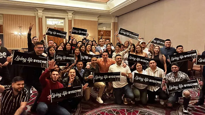 Cristoffer Serrano celebrating his 1-Star President achievement with the ByDzyne team, holding "Living Life ByDzyne" banners at a recognition event.