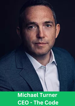 A professional headshot of Michael Turner, CEO of The Code, wearing a dark blue suit with a confident expression against a dark background.