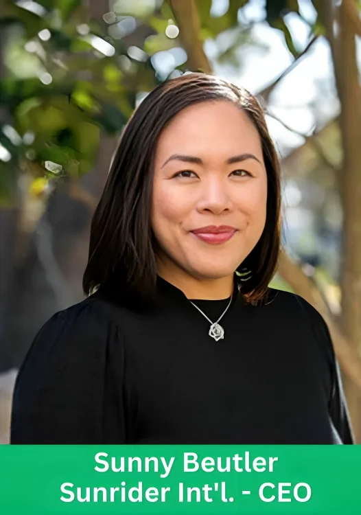 Sunny Beutler, CEO of Sunrider International, smiling with a necklace, standing against a natural background.
