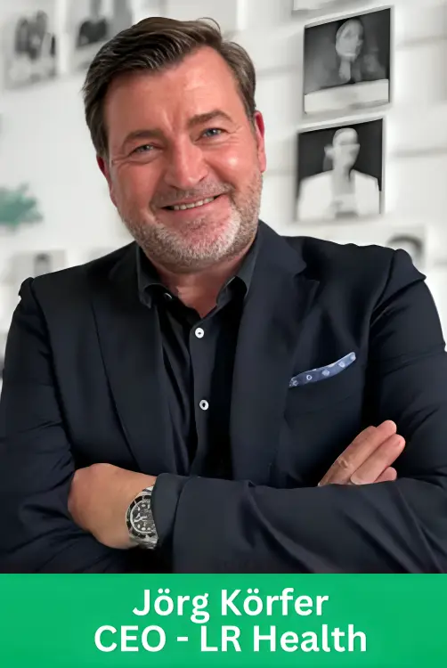 Professional corporate portrait of Jörg Körfer, CEO of LR Health, wearing a dark suit and black shirt with arms crossed, standing in front of a white wall with framed pictures.